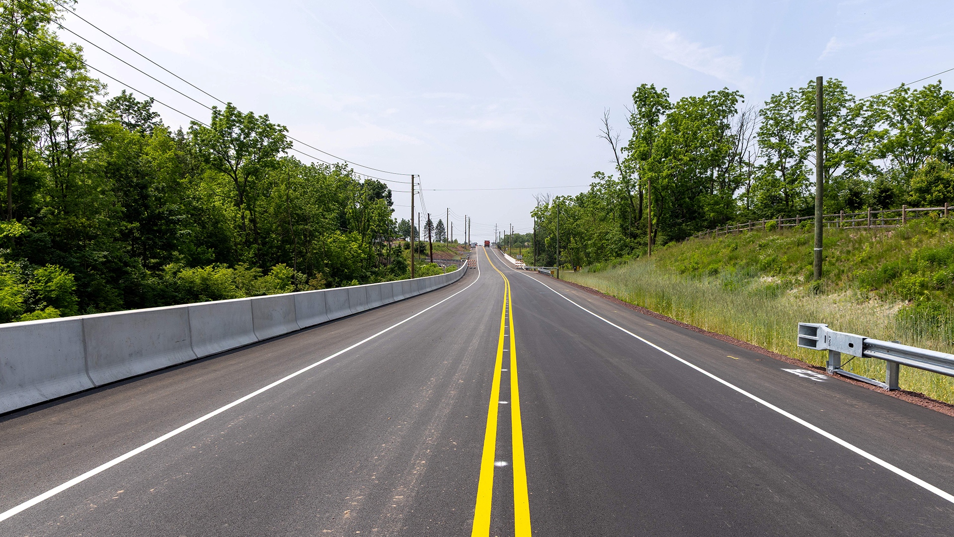 A recently paved and painted two-lane road with light woods on both sides and a waist-height concrete barrier on one side.